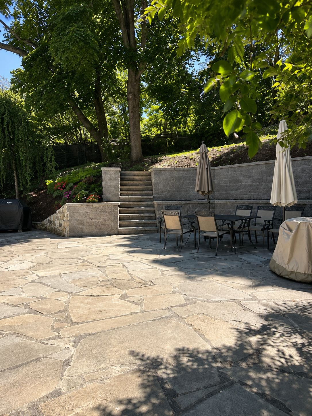 Flagstone patio with a stone retaining wall, steps, and outdoor dining set beneath mature trees, a Truestone outdoor living project